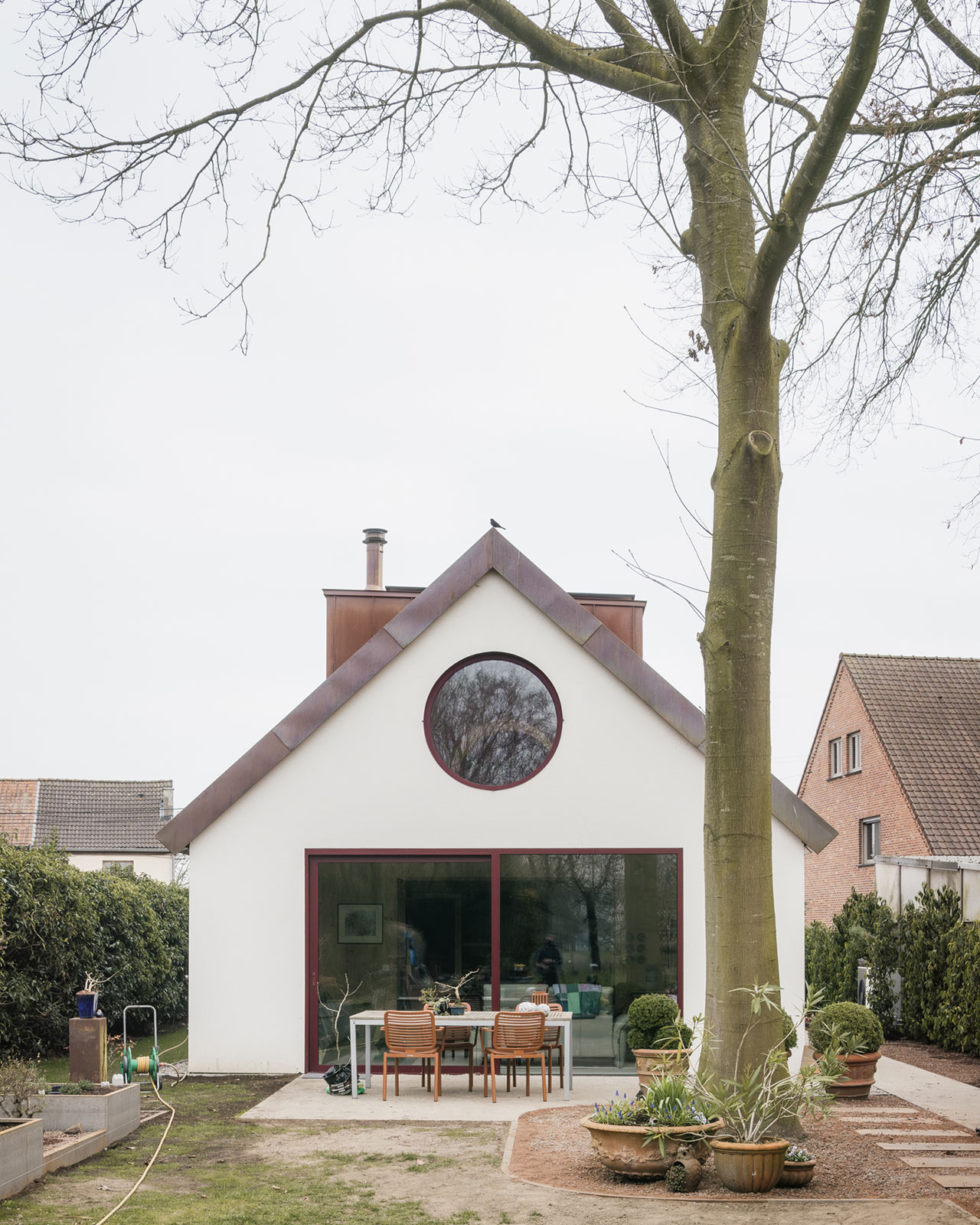 Modern white house with a large round window above a glass sliding door, FELT roofing accents, outdoor dining table and chairs, and a tall tree in the foreground.