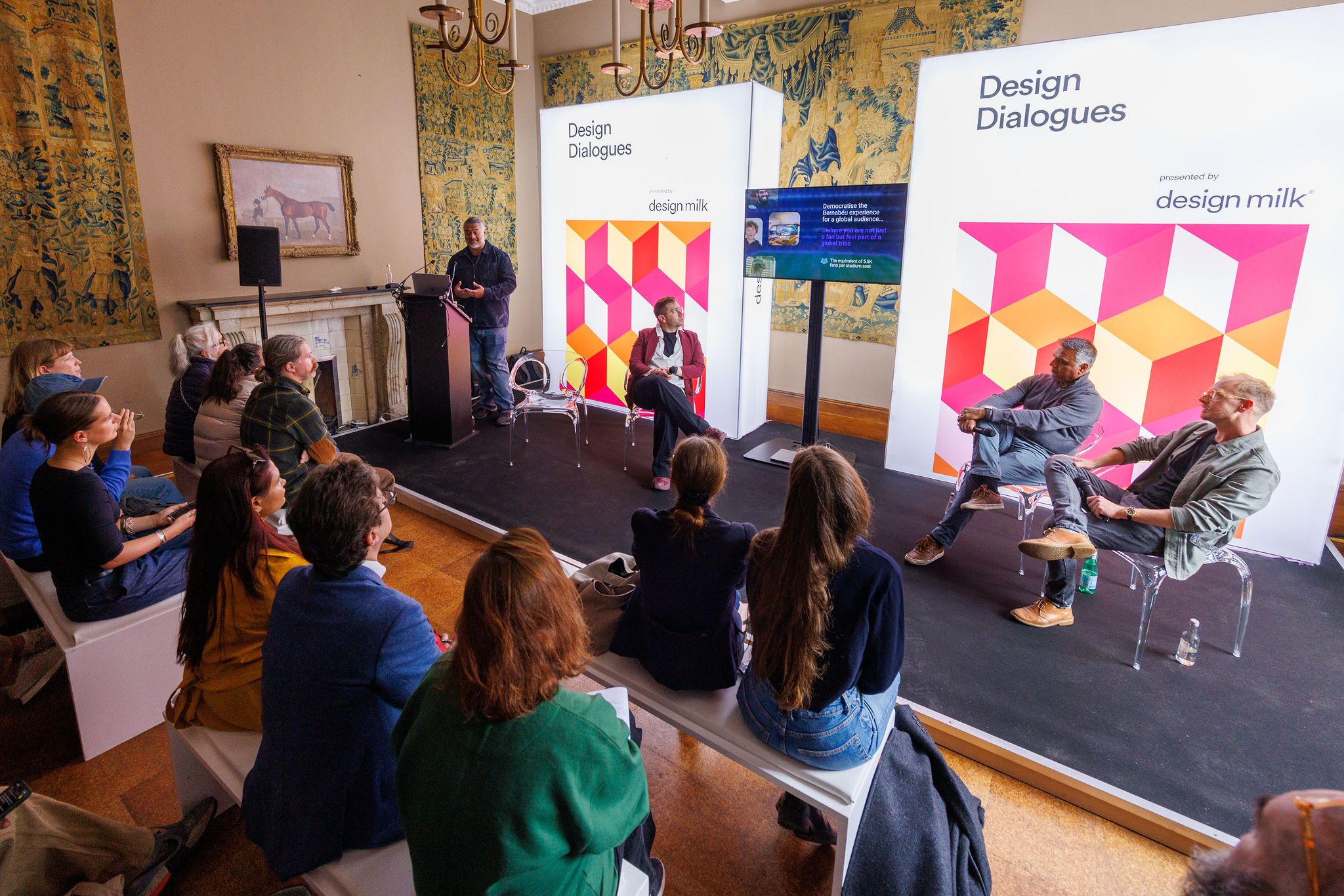 A seated audience listens to a panel of four speakers discussing design on stage beneath 