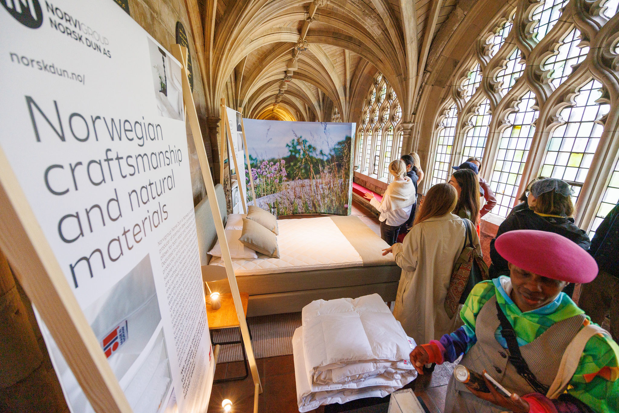 Visitors observe an exhibit at Clerkenwell Design Week, featuring Norwegian craftsmanship and natural materials, with a bed display set up in a historic, arched corridor with large windows.