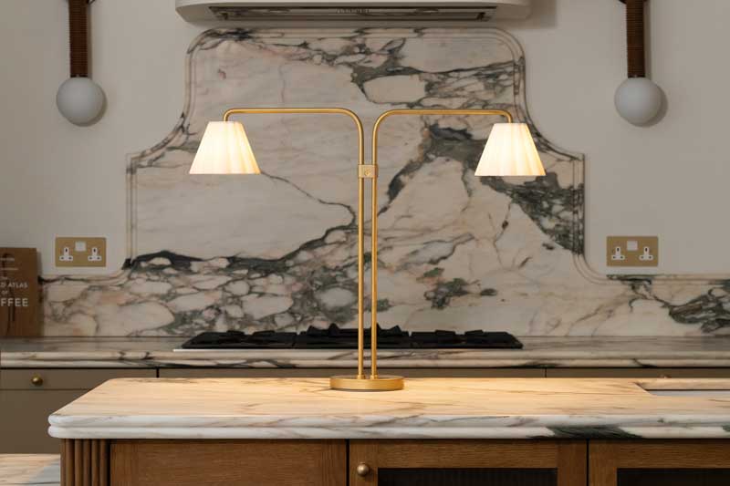 A brass double-shaded table lamp sits on a marble kitchen island, echoing the modern elegance showcased at Clerkenwell Design Week, with a matching marble backsplash and stove in the background.