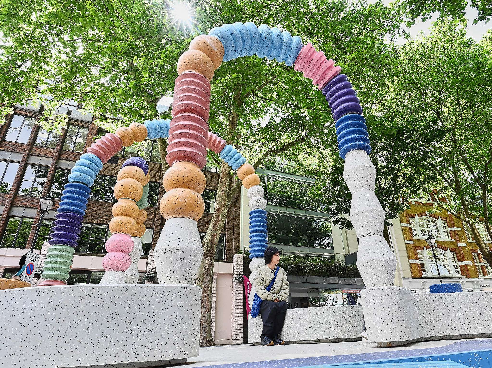 A person sits on a speckled concrete bench under colorful arched sculptures in an outdoor urban area with trees and modern buildings in the background.
