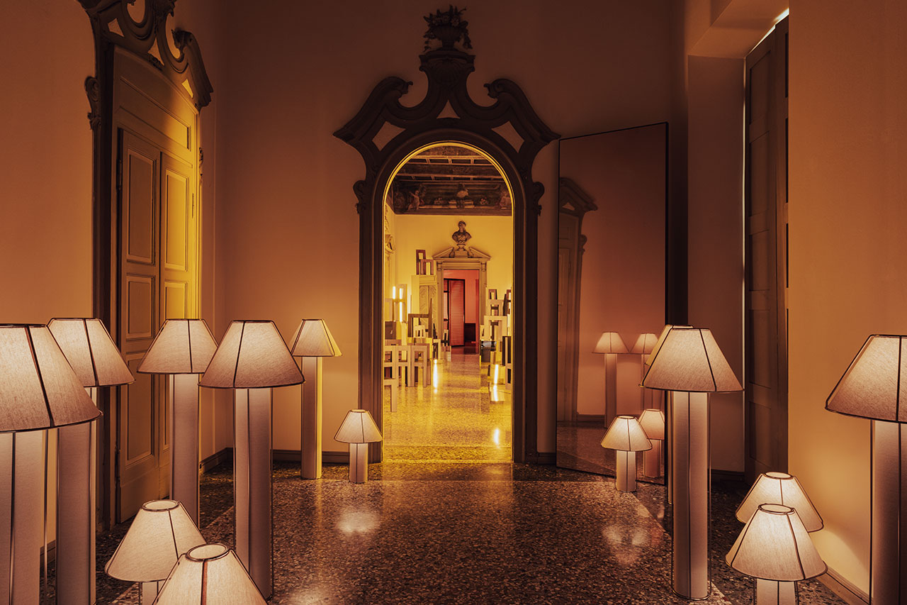 A hallway with ornate doorways and polished floors is lit by numerous standing lamps with beige shades, leading to a room with shelves and more lamps.