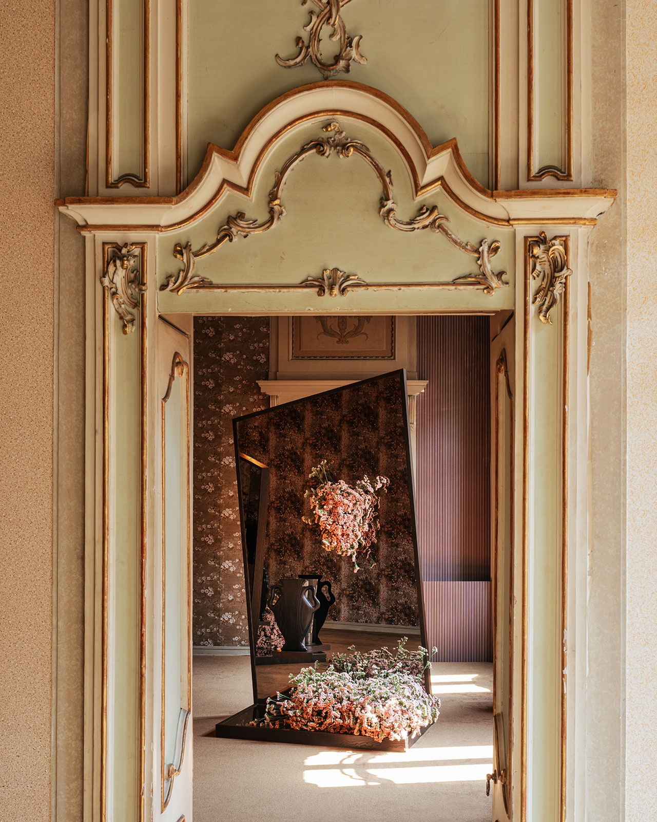 A view through ornate double doors reveals a modern floral art installation with mirrors and flowers inside a richly decorated room.