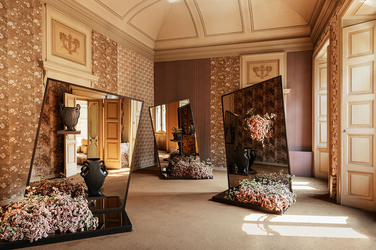 A decorative room with floral wallpaper, large angled mirrors reflecting vases filled with pink flowers, and sunlight coming through tall windows.