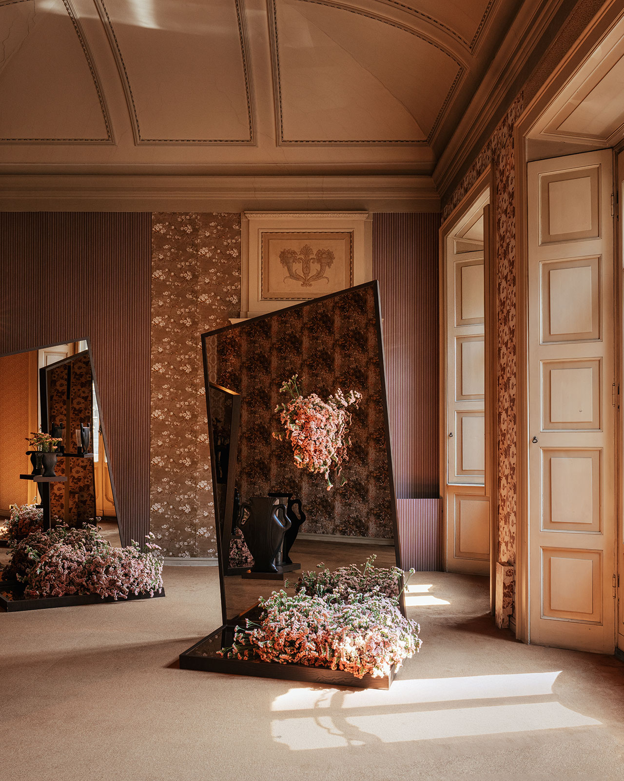 A large mirror stands at an angle in a decorated room with floral arrangements on the floor and reflected in the mirror, next to tall windows letting in sunlight.