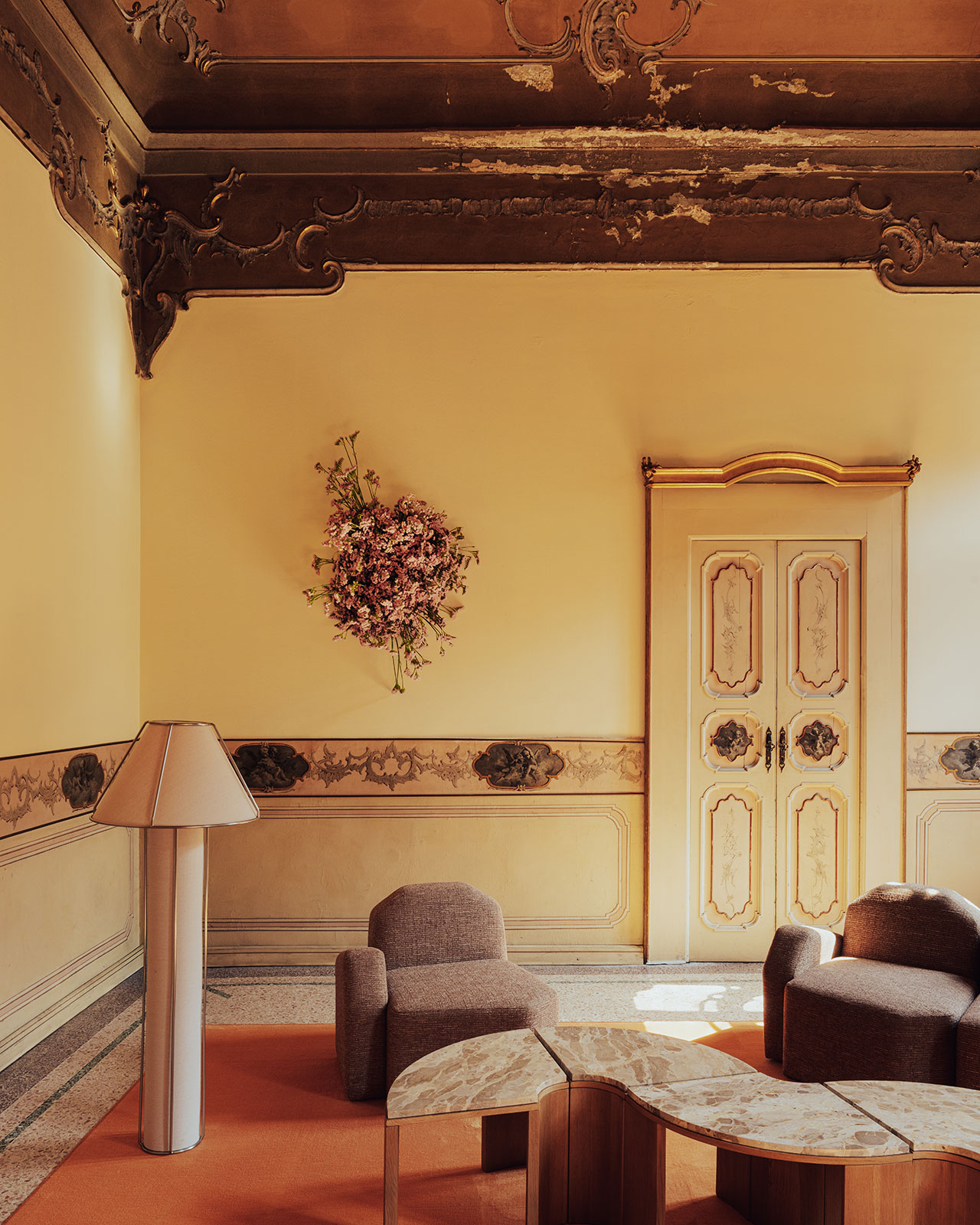 Elegant room with ornate moldings, two armchairs, a geometric marble coffee table, a tall modern lamp, a wall-mounted floral arrangement, and a decorative door.