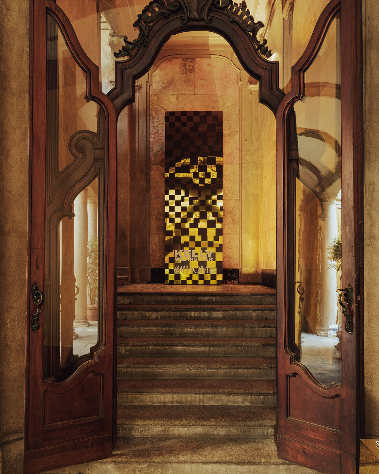 A grand wooden doorway opens to stone steps leading up to a hall with a tall, checkered yellow and brown modern art installation at the landing.