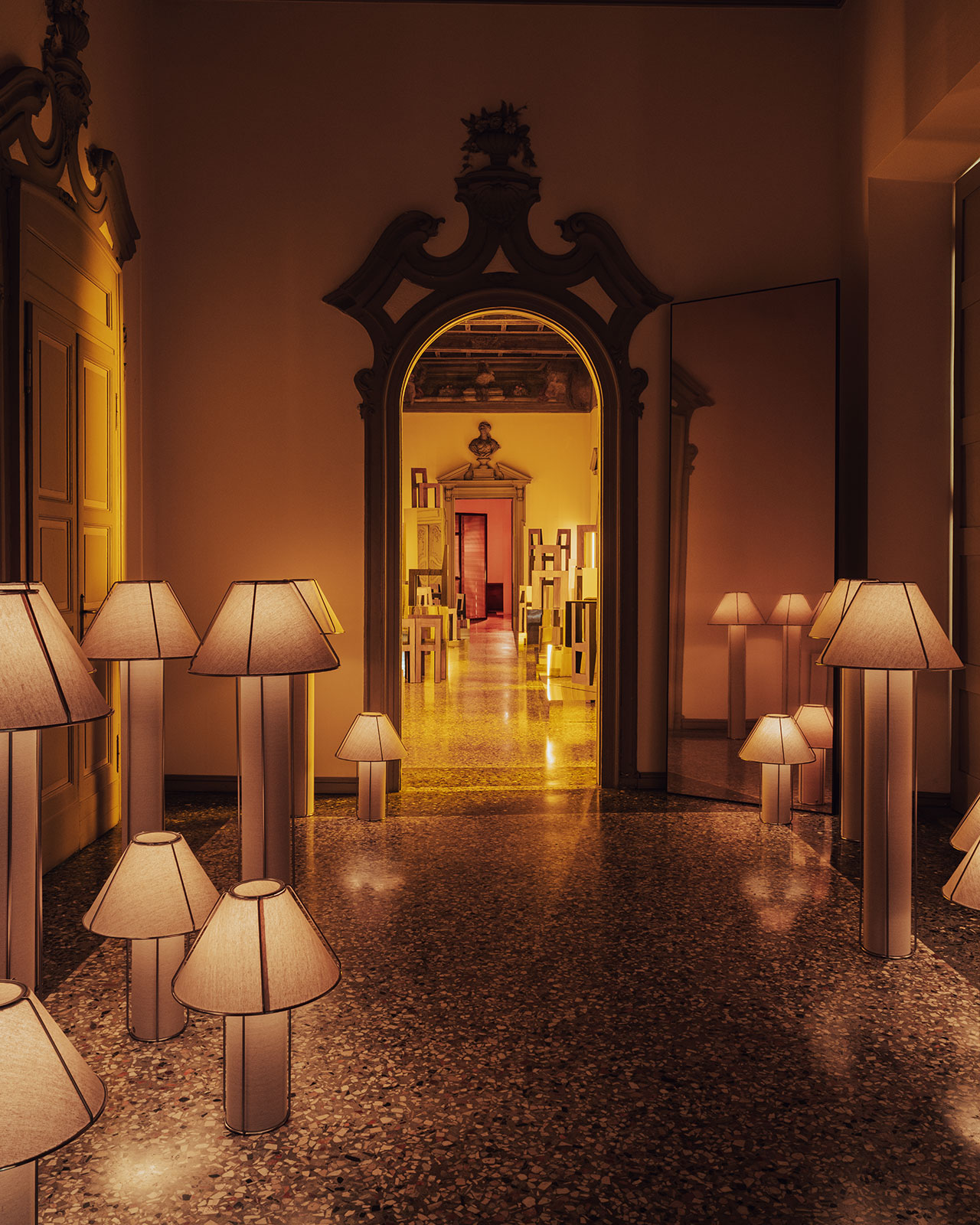 A hallway with ornate doorways features numerous floor and table lamps with beige shades, casting a warm yellow light on polished stone floors.