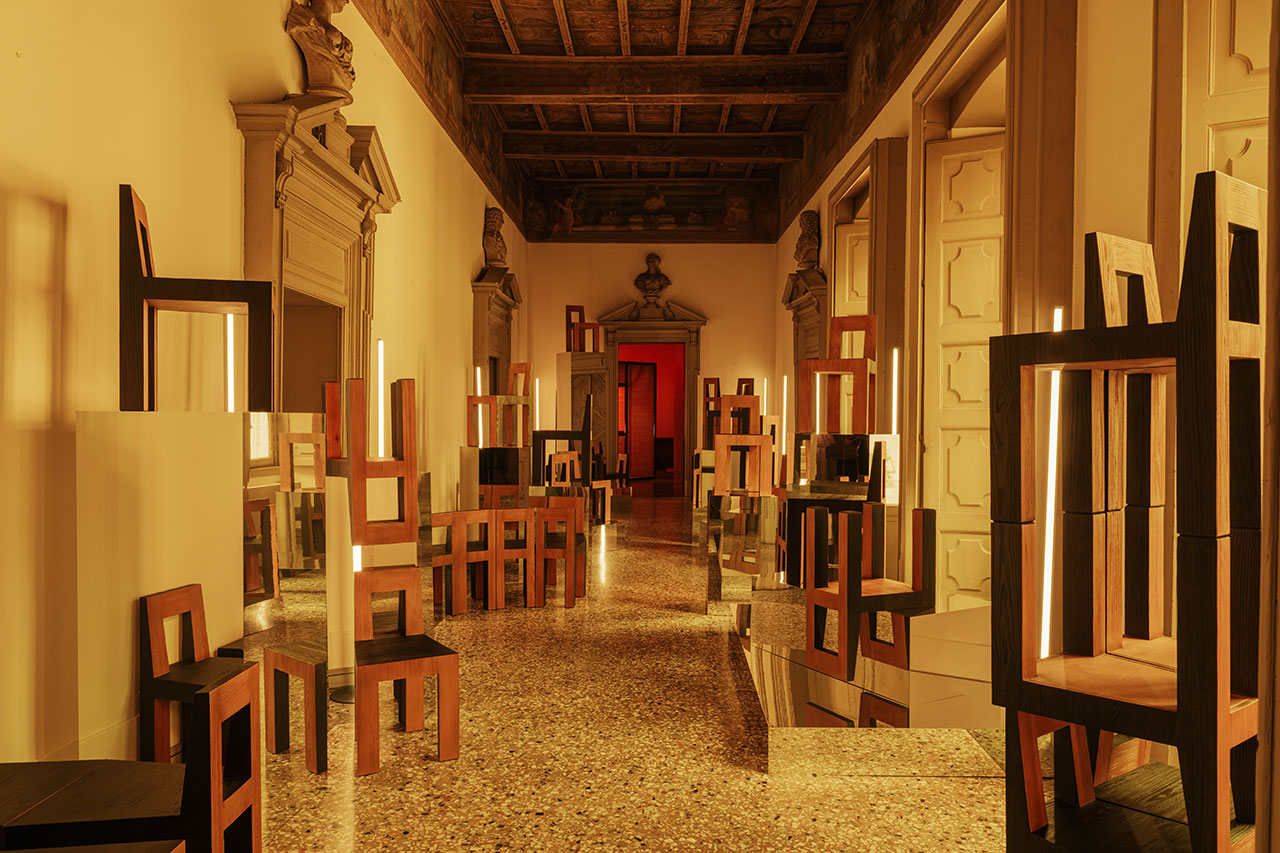 A hallway with polished stone floors, decorated with stacked wooden chairs and vertical light strips, featuring ornate door frames and a wooden coffered ceiling.