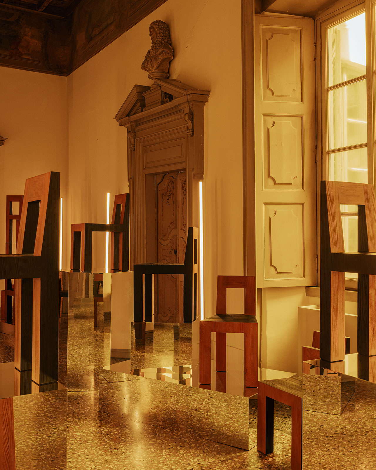 A room with classical architecture, large windows, and modern geometric wooden chairs reflected in mirrored surfaces on the floor.
