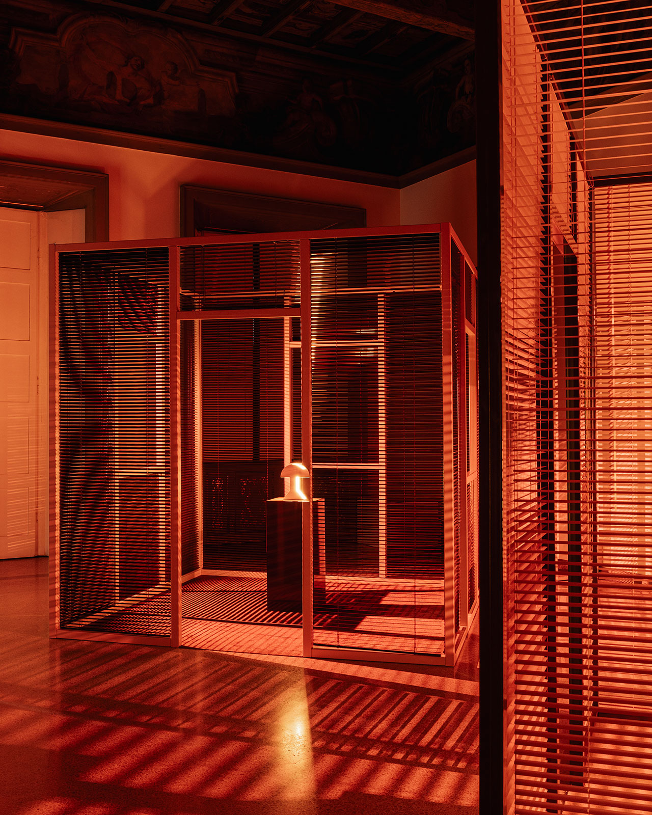 Sculptural installation with wooden slatted walls and orange lighting, featuring a small object on a pedestal inside a cubic structure.