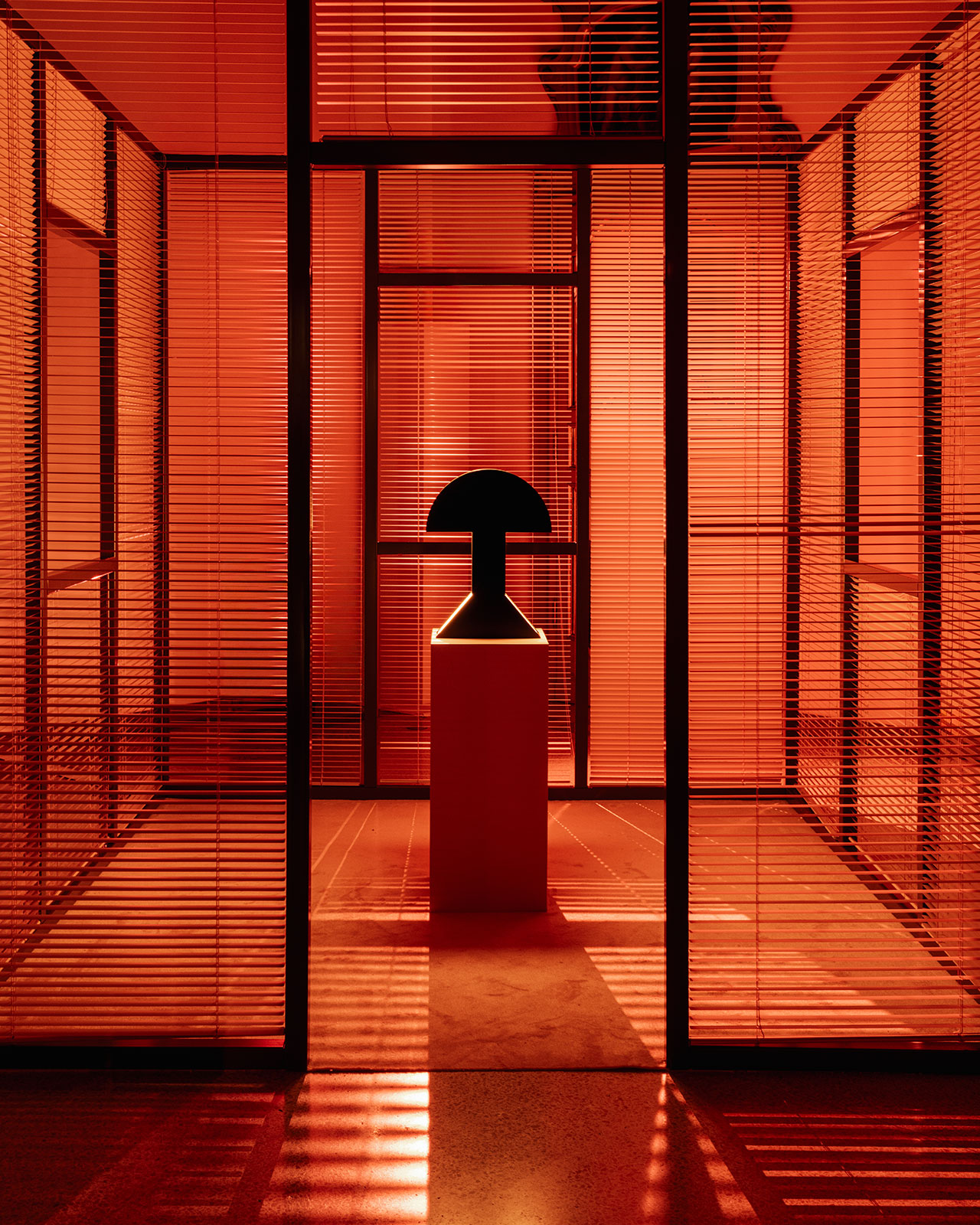 A dark geometric sculpture on a pedestal is centered in a room bathed in red light, viewed through glass panels with horizontal blinds.