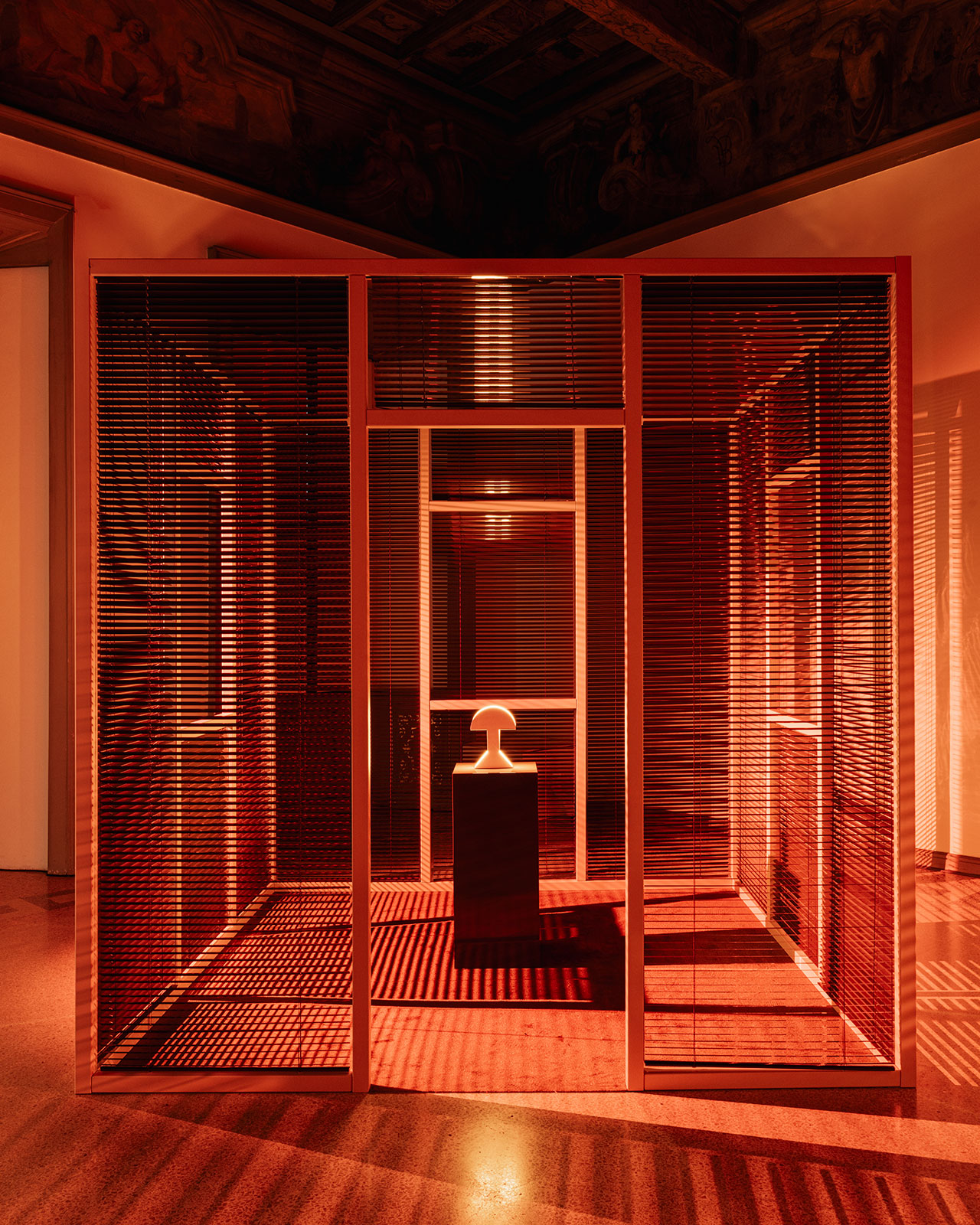 A small sculpture on a pedestal is displayed inside a geometric room with red lighting and slatted walls, casting linear shadows on the floor.