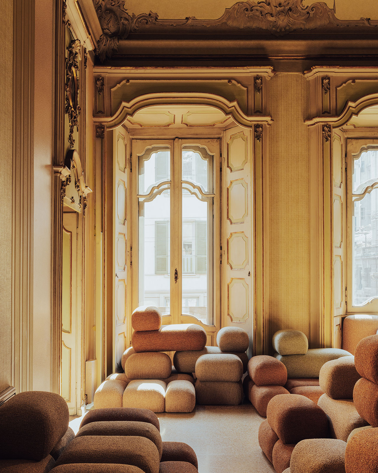 Ornate room with tall windows, detailed moldings, and clusters of earth-toned, modular seating arranged along the floor and under the windows.