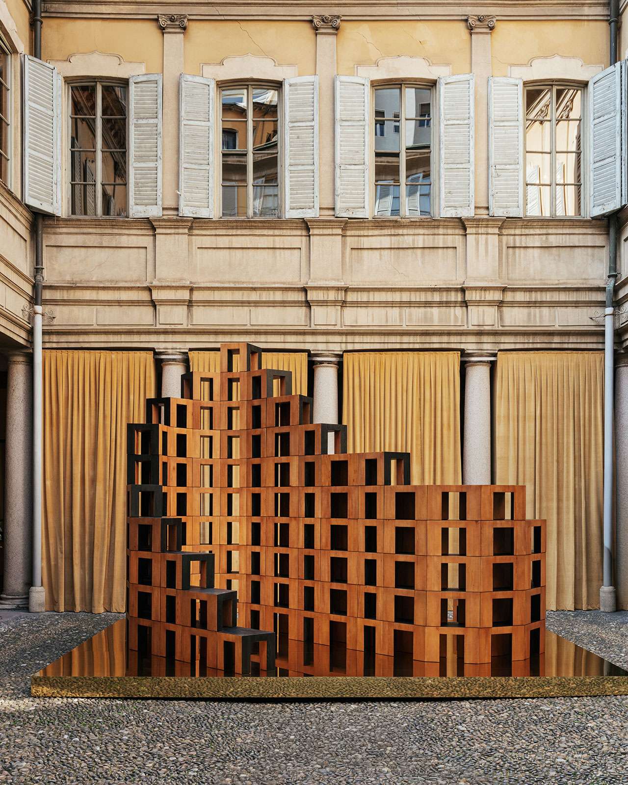 A geometric wooden sculpture with a stepped, grid-like pattern stands in a courtyard with stone columns and tan curtains, set against a historic building with shuttered windows.