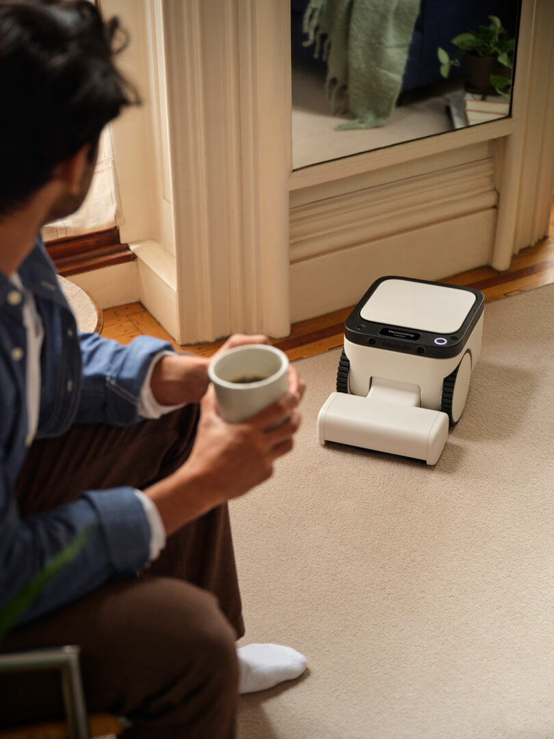 A person holding a mug sits on a carpeted floor while a small, white, box-shaped robot is positioned nearby.
