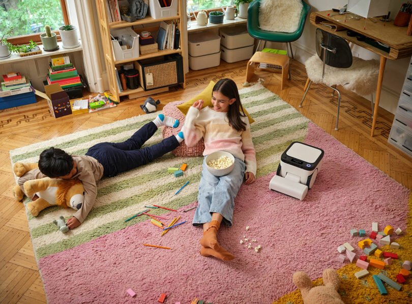 Two children sit and lie on a colorful rug in a cozy, cluttered room with toys, books, and a small robot. One child eats popcorn, and the other hugs a large stuffed animal.