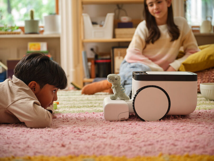 A child lies on a pink and white rug facing a small robot with a toy dinosaur on top, while another child sits in the background.