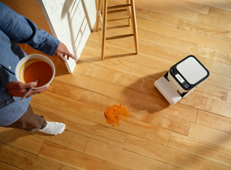 A person holding a bowl of soup points at a spill on a wooden floor while a robot mop approaches the mess.