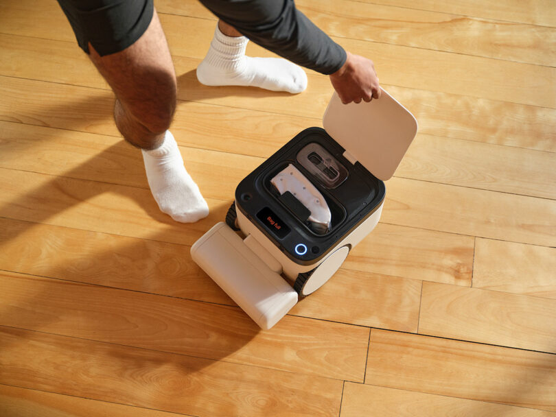 A person in white socks and shorts is opening the lid of a small robotic cleaning device on a wooden floor.