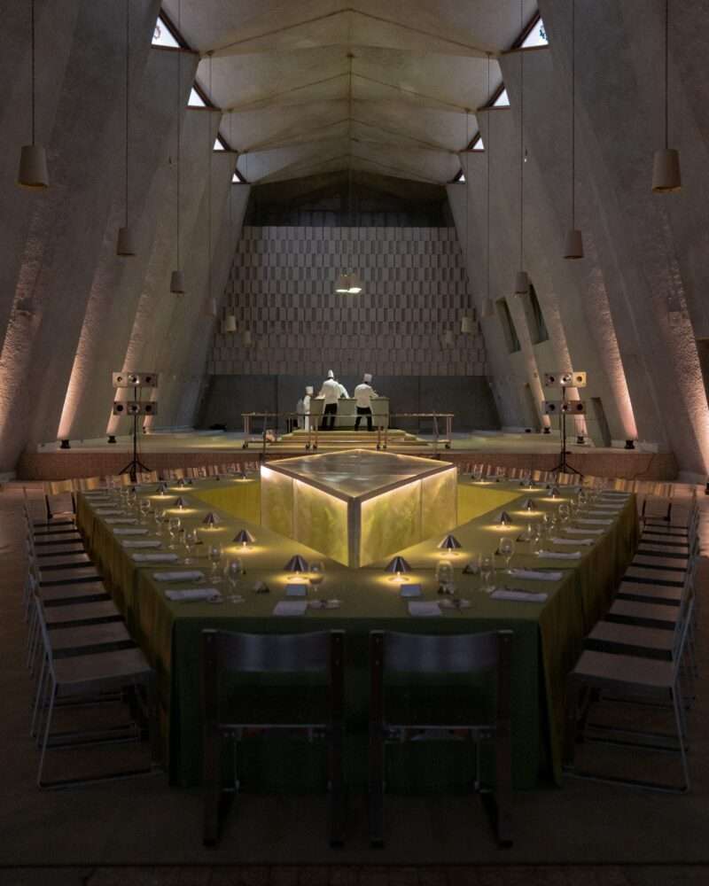 A long, triangular dining table with place settings and a green tablecloth is set in a modern, high-ceilinged hall; two people stand at the far end behind a counter featuring a Menu Extra display.