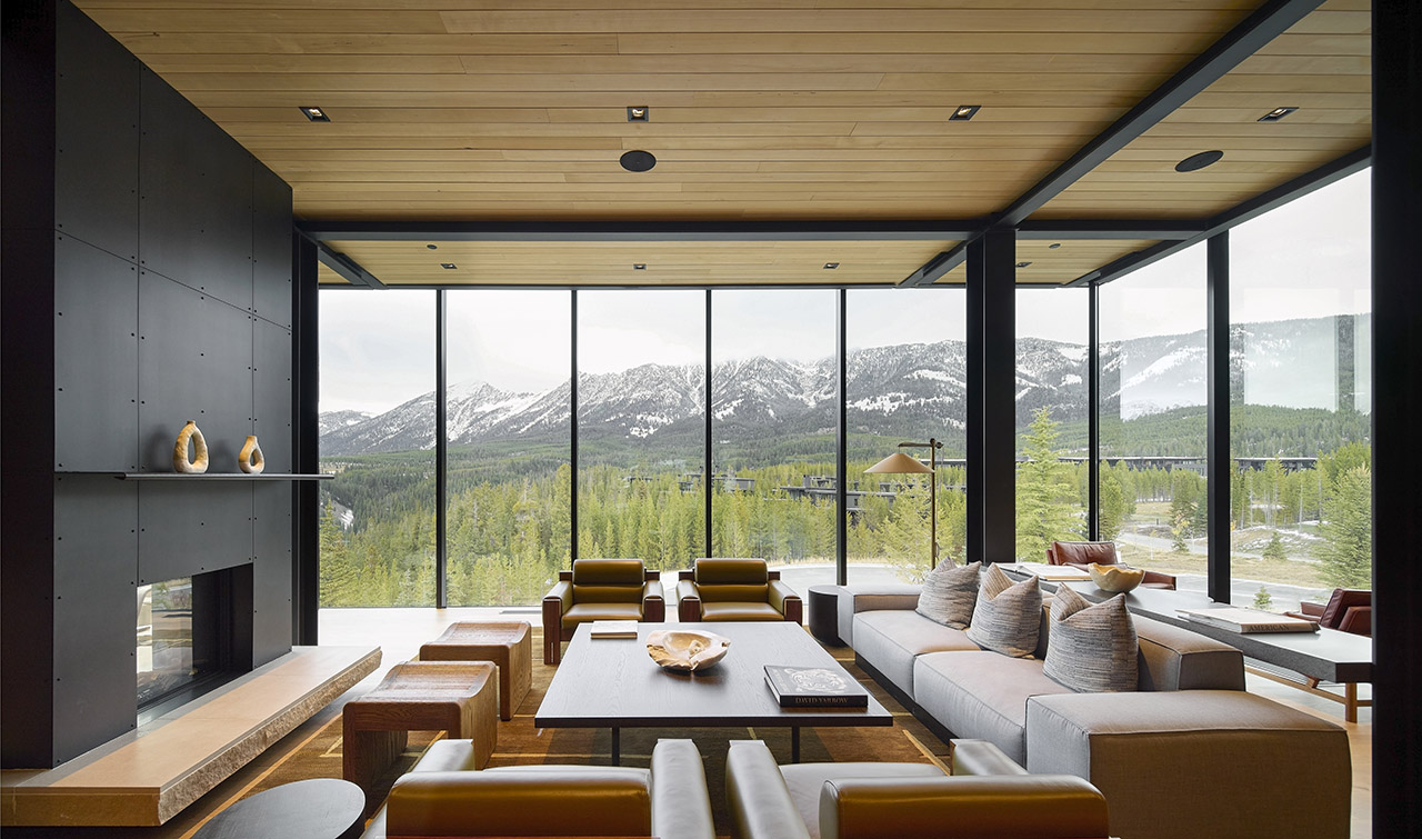Modern living room with large floor-to-ceiling windows offering a panoramic view of snow-capped mountains and pine trees. Neutral, minimalist furnishings and a black fireplace complete the space.