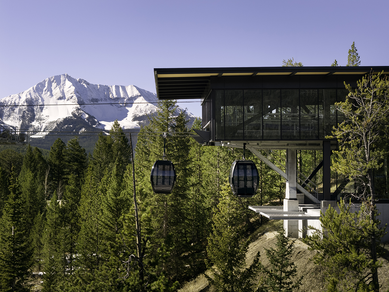 A modern gondola lift station with two gondolas is set among pine trees, with snow-capped mountains visible in the background under a clear sky.