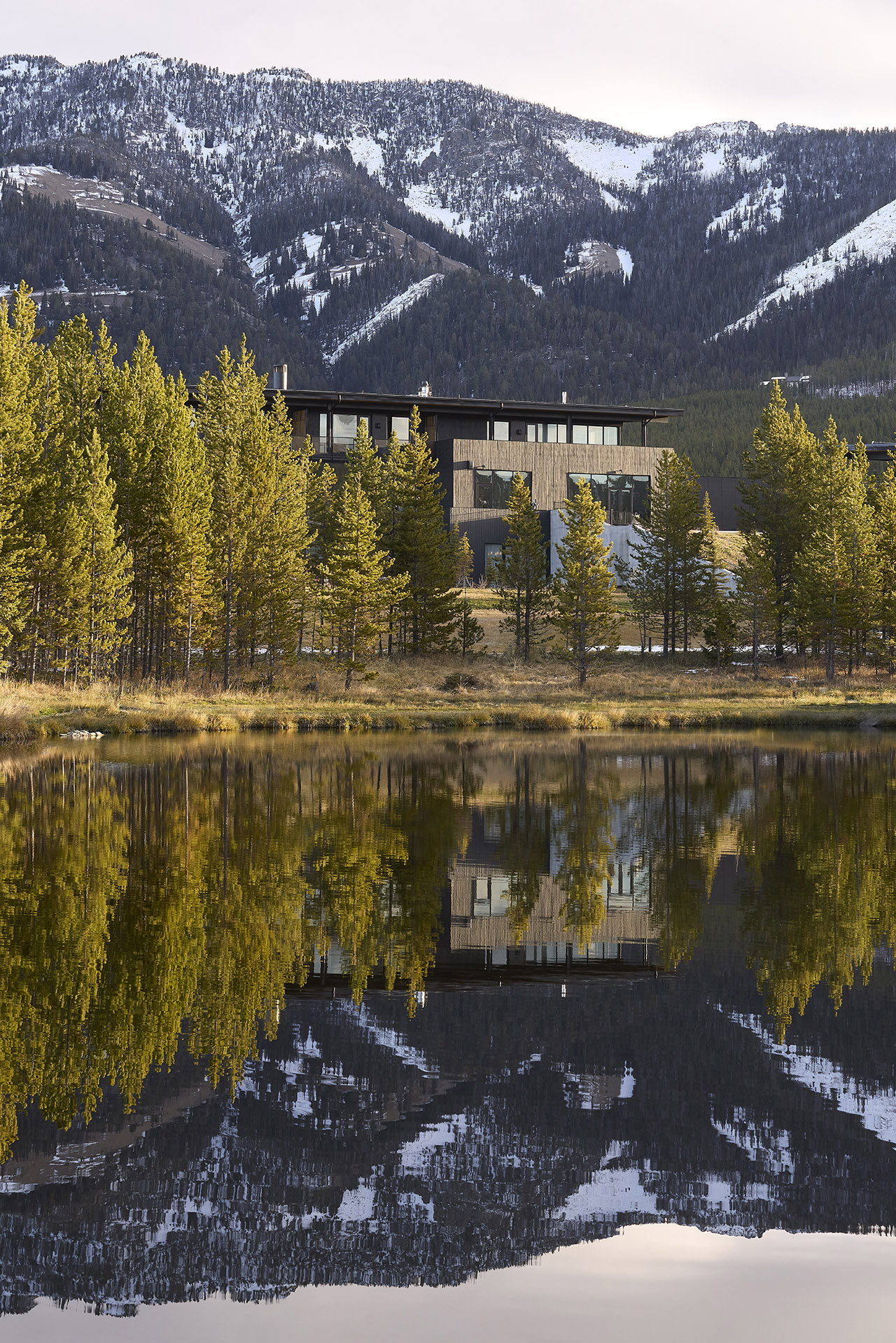 Modern house with large windows surrounded by pine trees, reflected in a calm lake, with snow-capped mountains in the background.