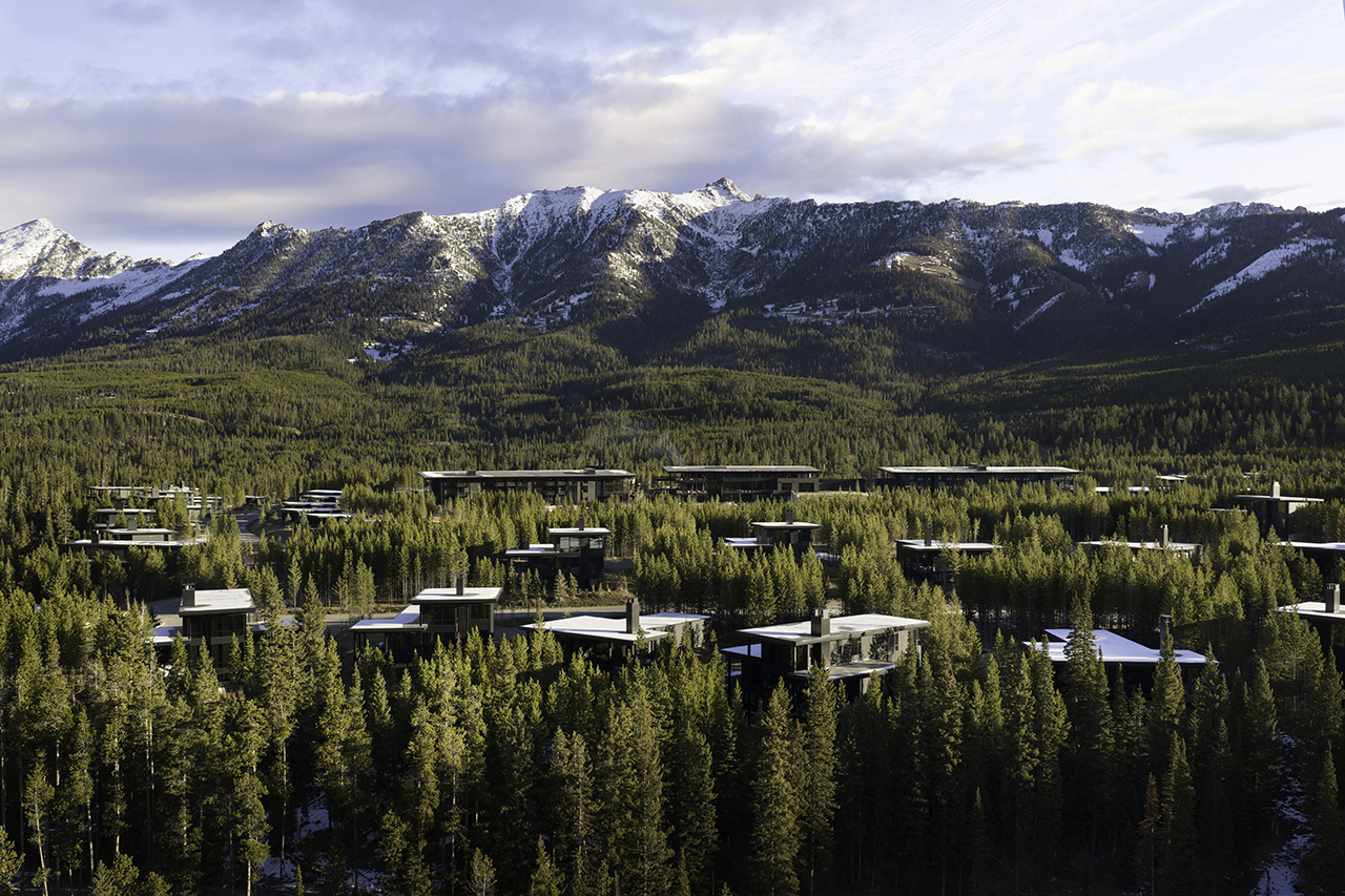 A cluster of modern, flat-roofed buildings surrounded by dense pine trees with snow-capped mountains in the background under a partly cloudy sky.