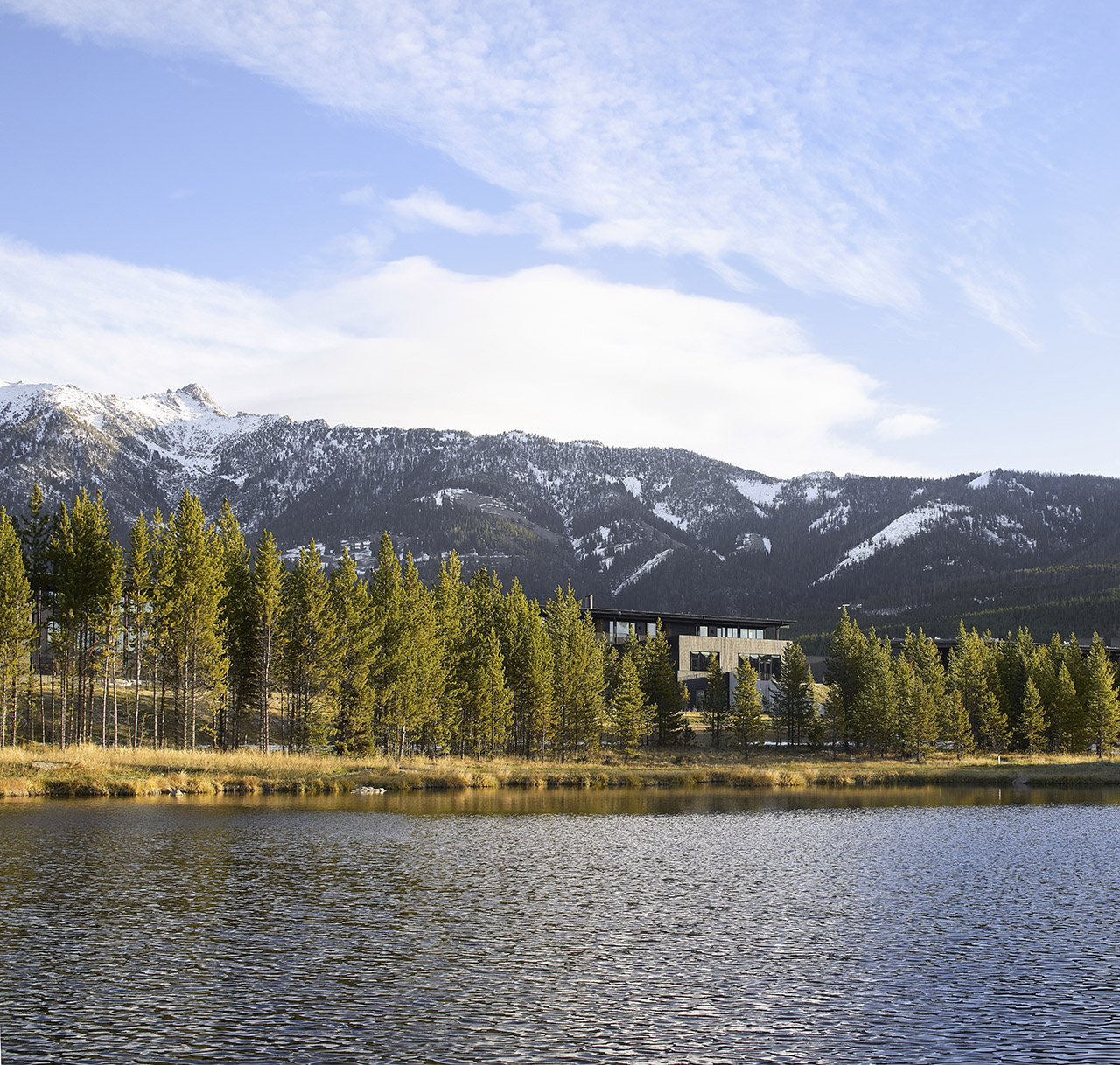 A modern building is situated among pine trees by a lake, with snow-capped mountains visible in the background under a partly cloudy sky.