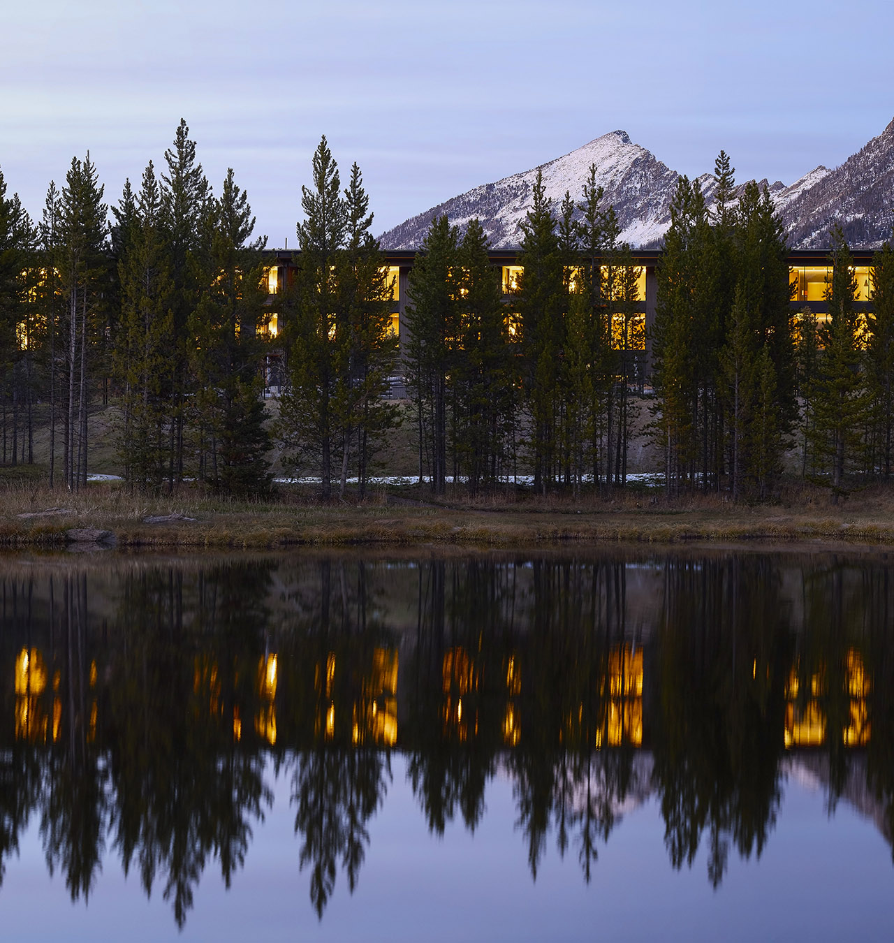 A modern building with illuminated windows is seen behind a row of pine trees, reflected in a calm lake, with a snow-capped mountain in the background at dusk.