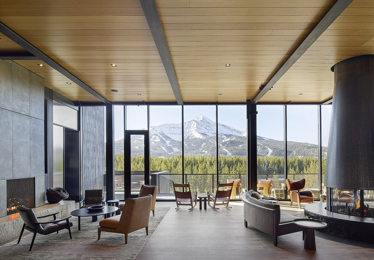 Modern lounge with large windows, contemporary furniture, and a fireplace, offering a scenic view of a snow-capped mountain and pine forest.