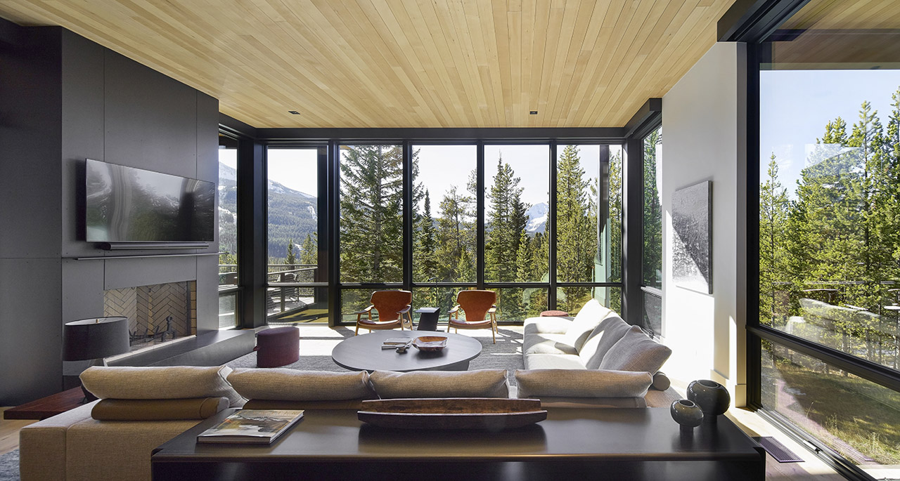 Modern living room with large floor-to-ceiling windows, mountain and forest views, light wood ceiling, neutral furniture, and a wall-mounted TV above a fireplace.