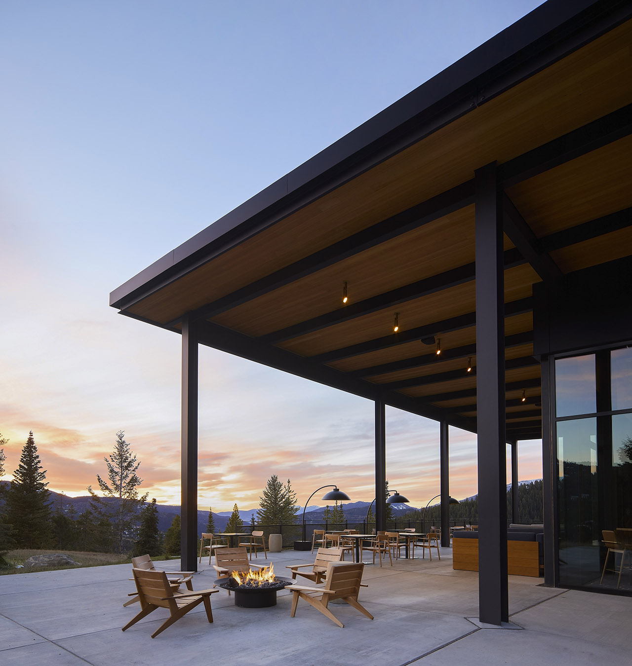 Covered outdoor patio with modern wooden furniture around a fire pit, overlooking trees and mountains at sunset.