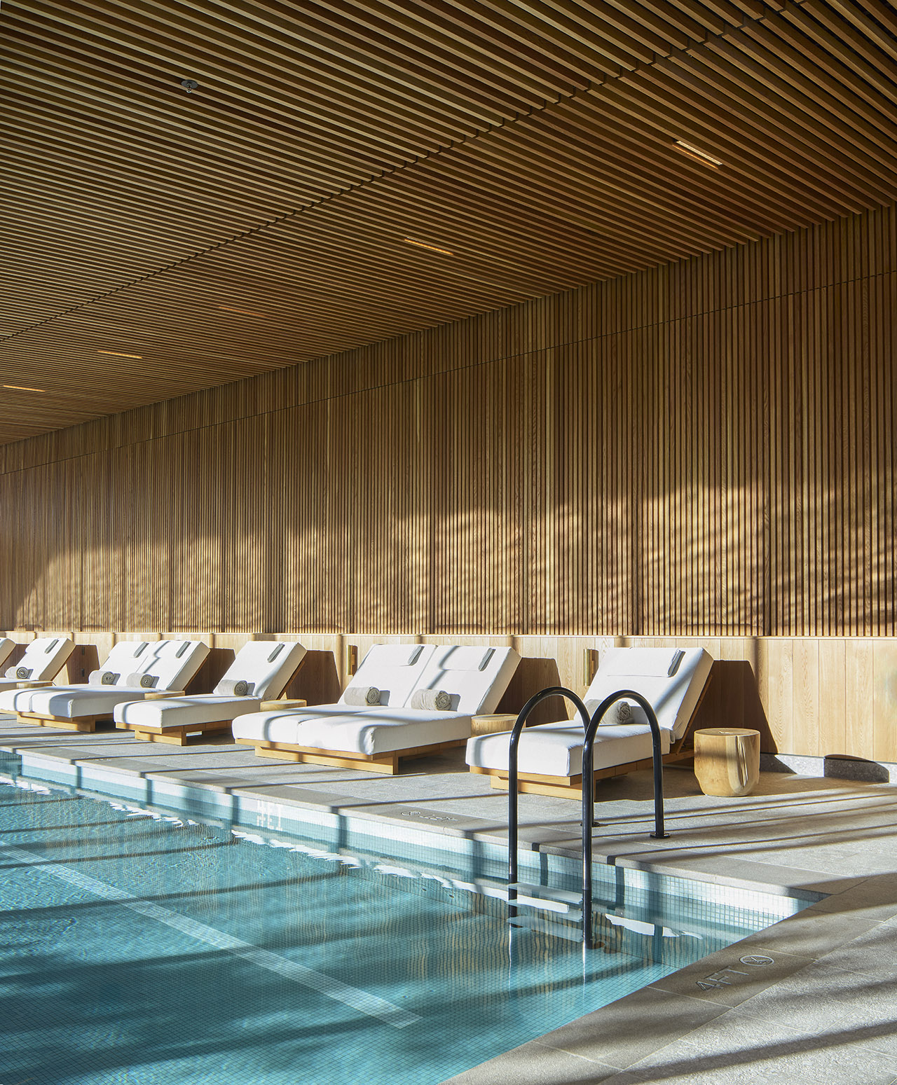 Indoor pool with clear water, several white cushioned lounge chairs, and a wooden slatted ceiling and wall, all illuminated by natural sunlight.