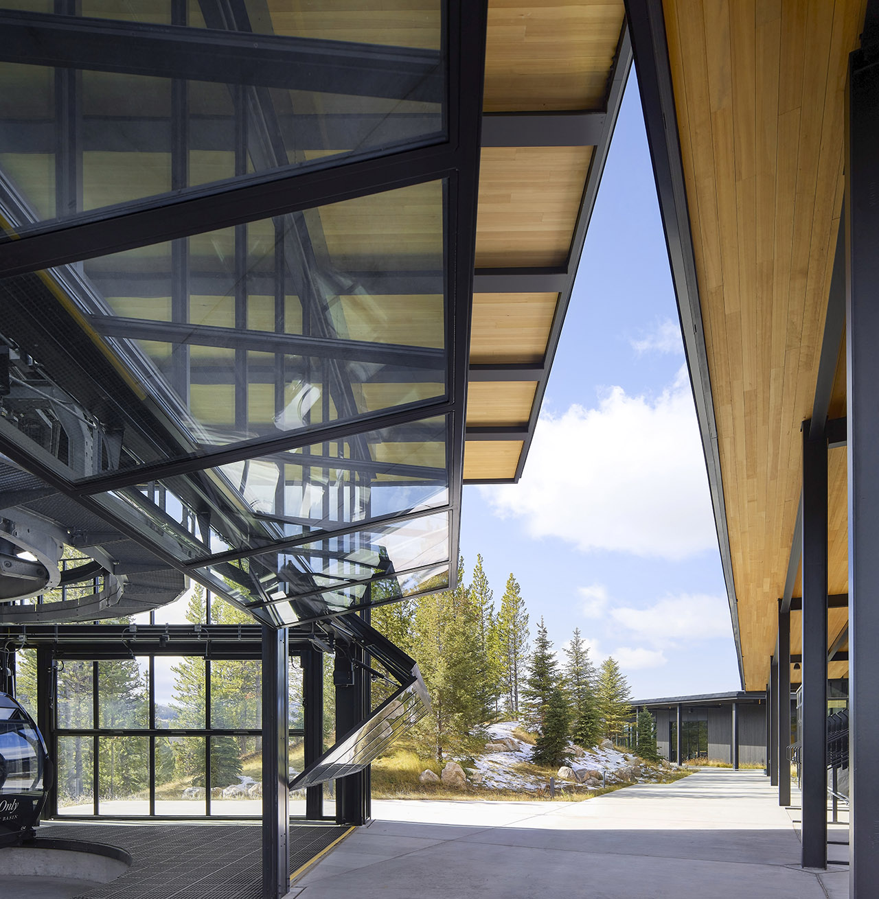 Modern glass and steel gondola station with angled roof, wooden ceiling panels, and outdoor area overlooking pine trees and a clear sky.