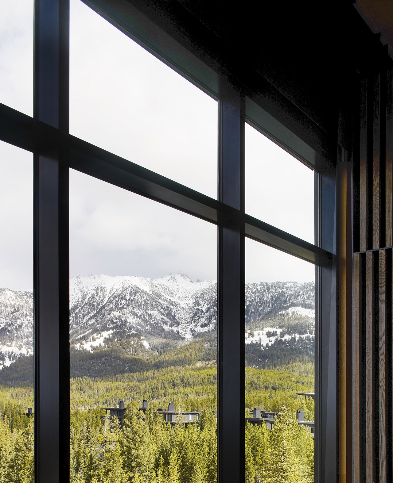 A view through large window panes shows snow-capped mountains and green pine trees under a cloudy sky, with rooftops visible below.