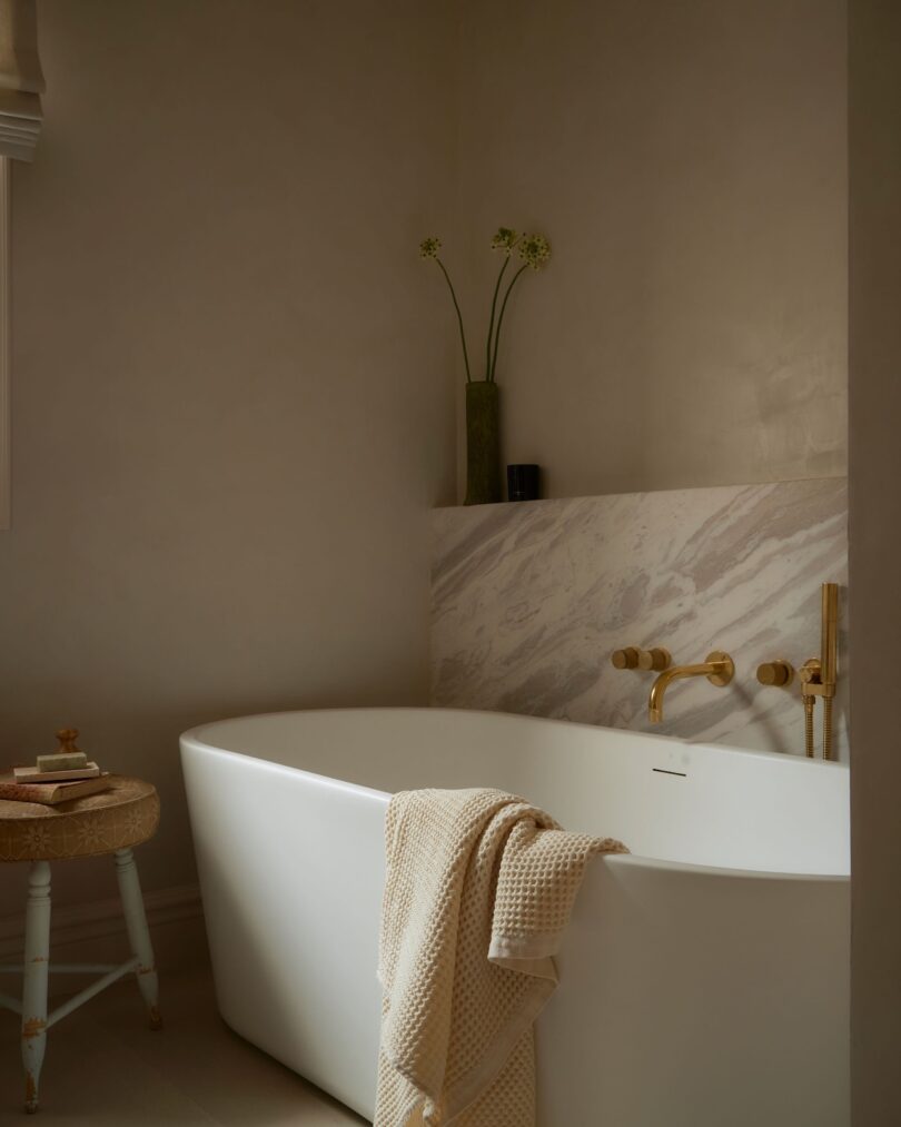 A modern bathroom with a freestanding white bathtub, beige towel draped over the side, marble backsplash, gold fixtures, and a vase with tall flowers on the ledge.