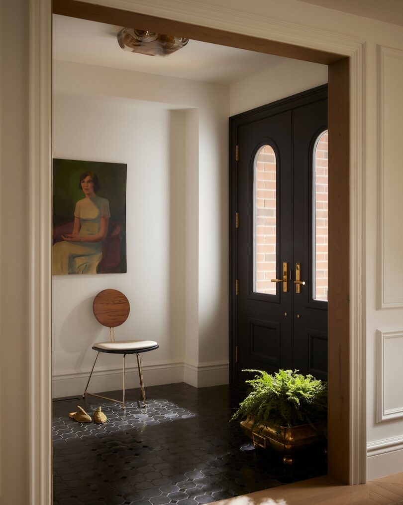 Entryway with a black double door, a fern in a gold planter, a round wooden chair, two duck figurines on the floor, and a portrait of a woman hanging on the wall.
