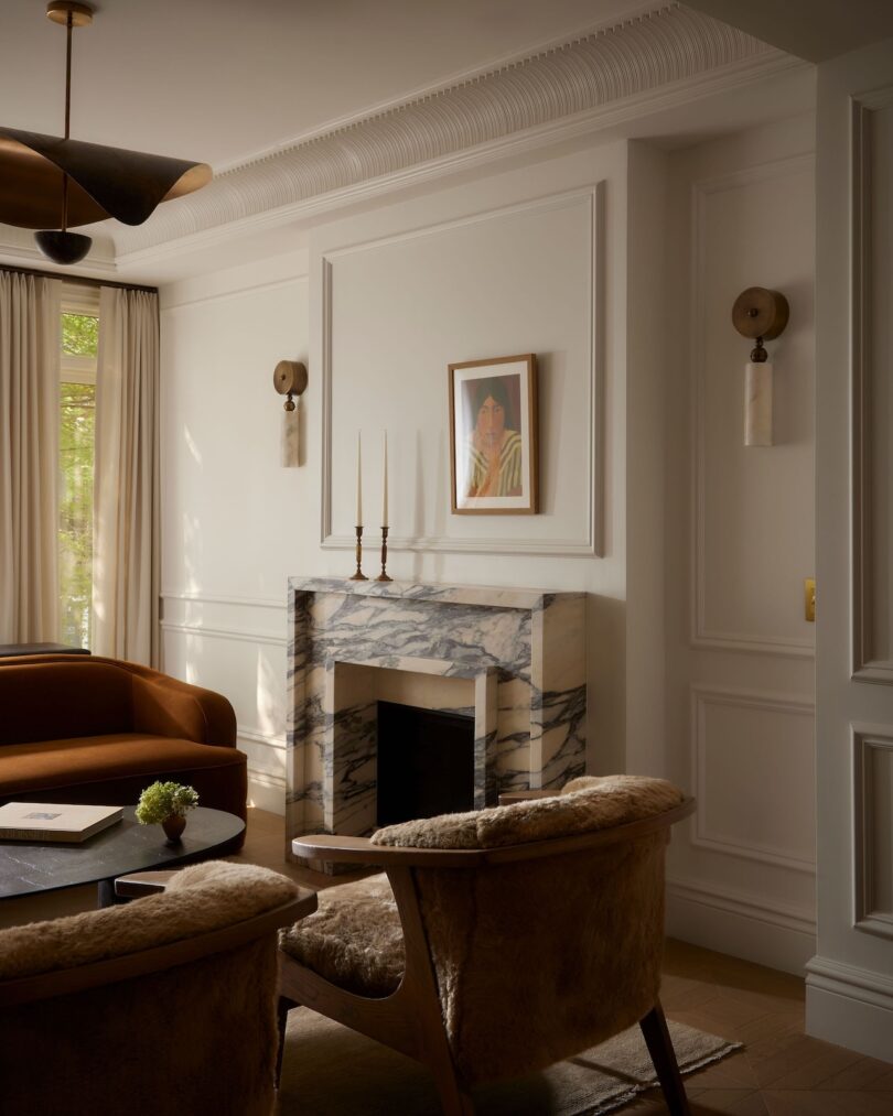 A stylish living room with a marble fireplace, two furry armchairs, a round coffee table, and a framed portrait above the mantel. Natural light enters through a window with curtains.