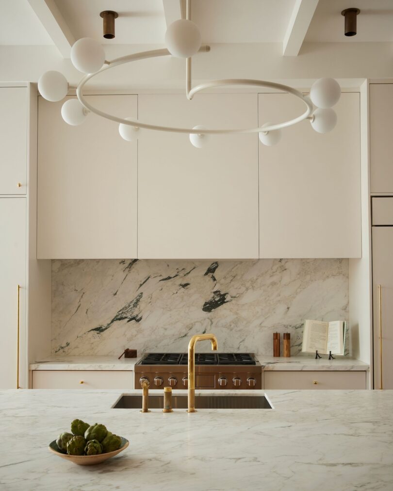 Modern kitchen with marble countertops and backsplash, gold faucet, large stove, built-in cabinets, circular light fixture, and a bowl of artichokes on the counter.