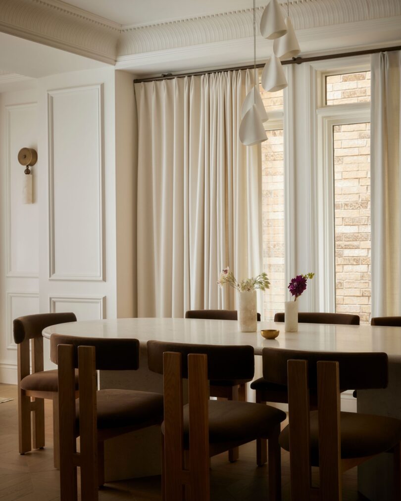 A modern dining room with a round table, dark cushioned wooden chairs, vases with flowers, beige curtains, pendant lights, and a window showing a brick wall outside.