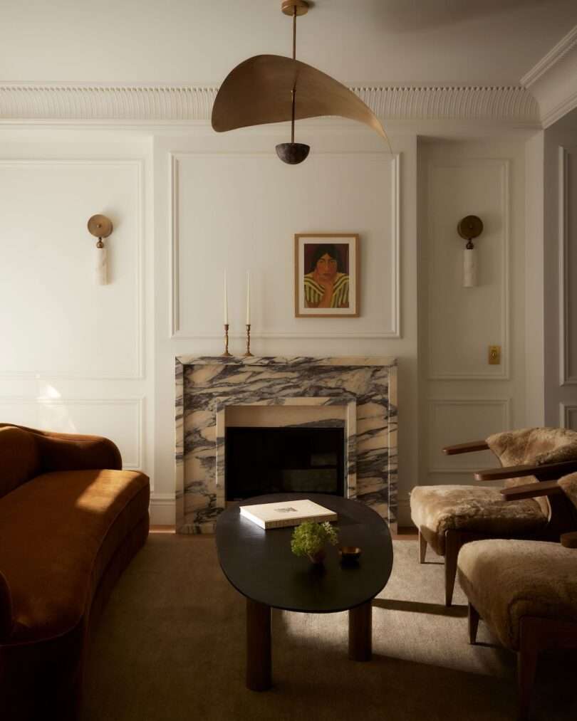 A living room with a marble fireplace, a modern black coffee table, brown sofa, armchair with fur cushion, wall sconces, and a portrait above the mantel.