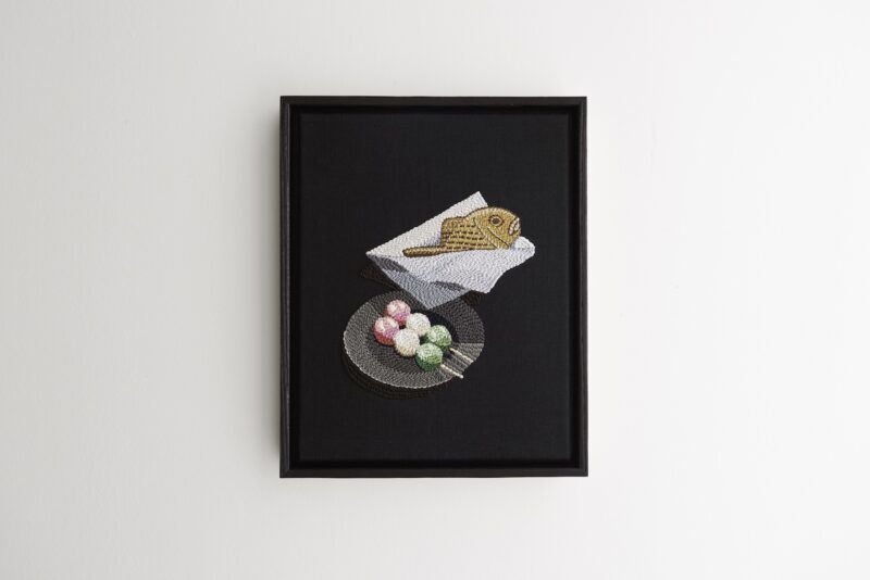 Framed art piece showing an embroidered taiyaki pastry in paper and a plate of colorful dango skewers on a black background.