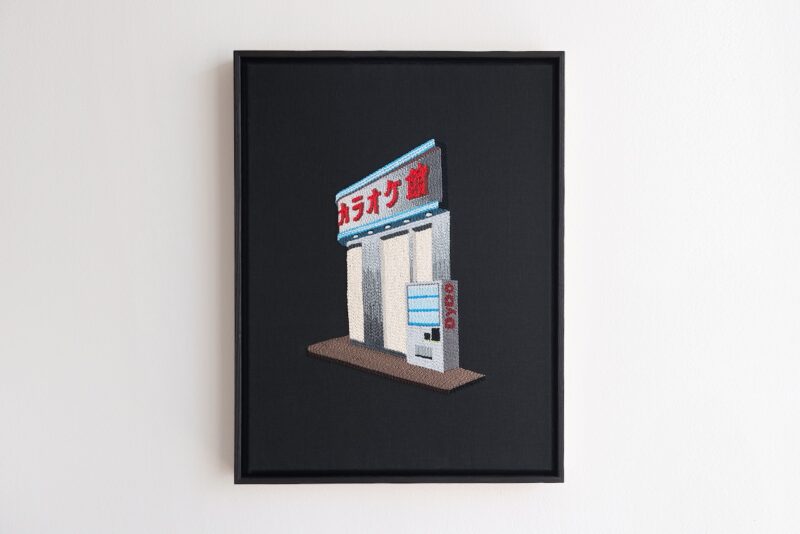 Framed embroidery of a small building with a vending machine and a sign in Japanese characters, displayed against a plain white wall.