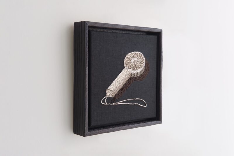 A framed textile artwork depicts a hair dryer, crafted from thread in shades of beige and brown, mounted on a black fabric background.