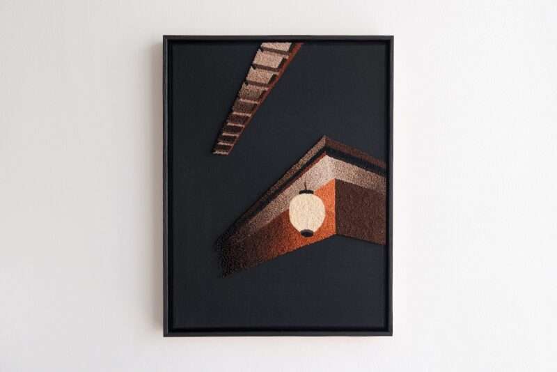Framed textile artwork depicting geometric roof shapes and a white lantern on a dark background, hanging on a white wall.