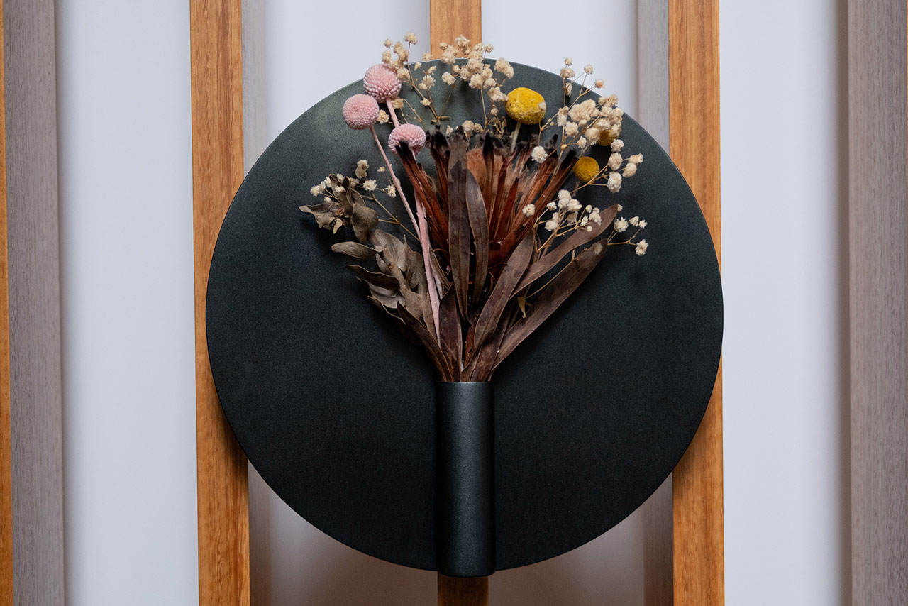 A black Nimbus Vase mounted on a round black plate holds dried flowers and foliage against a wooden slat background.