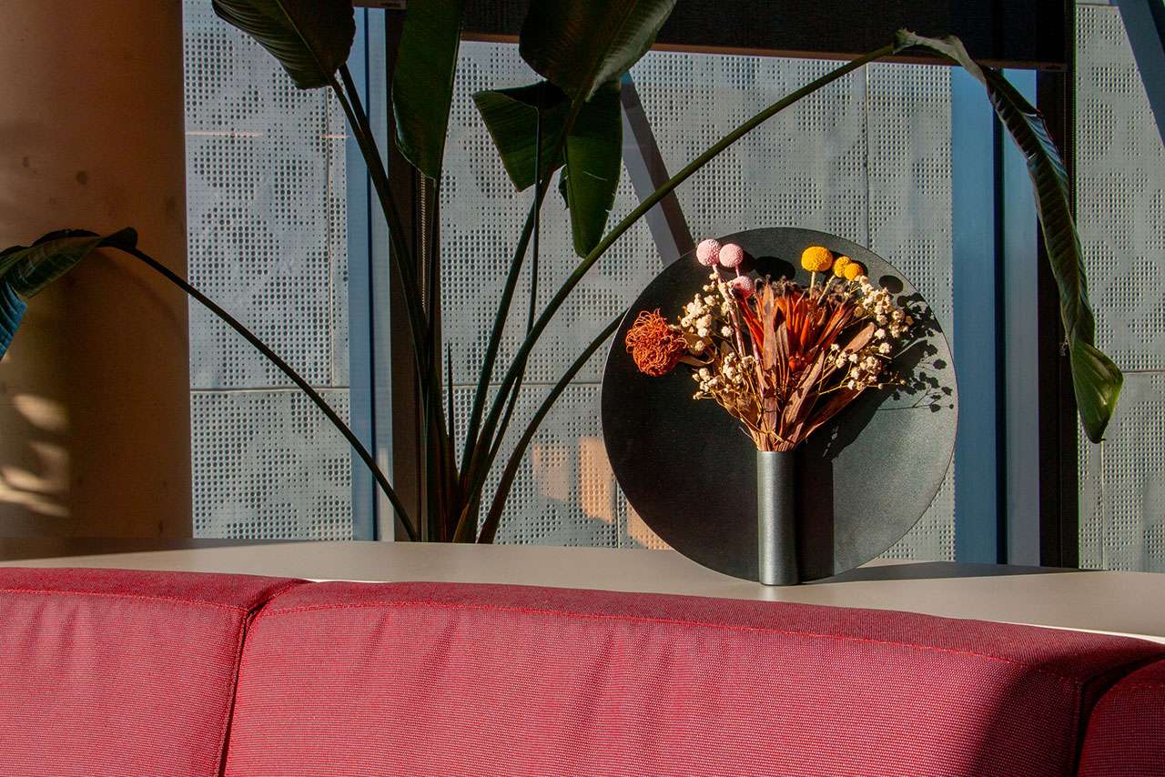 A Nimbus Vase with dried flowers sits on a white table behind a red couch, framed by large green plants and a window with perforated panels in the background.