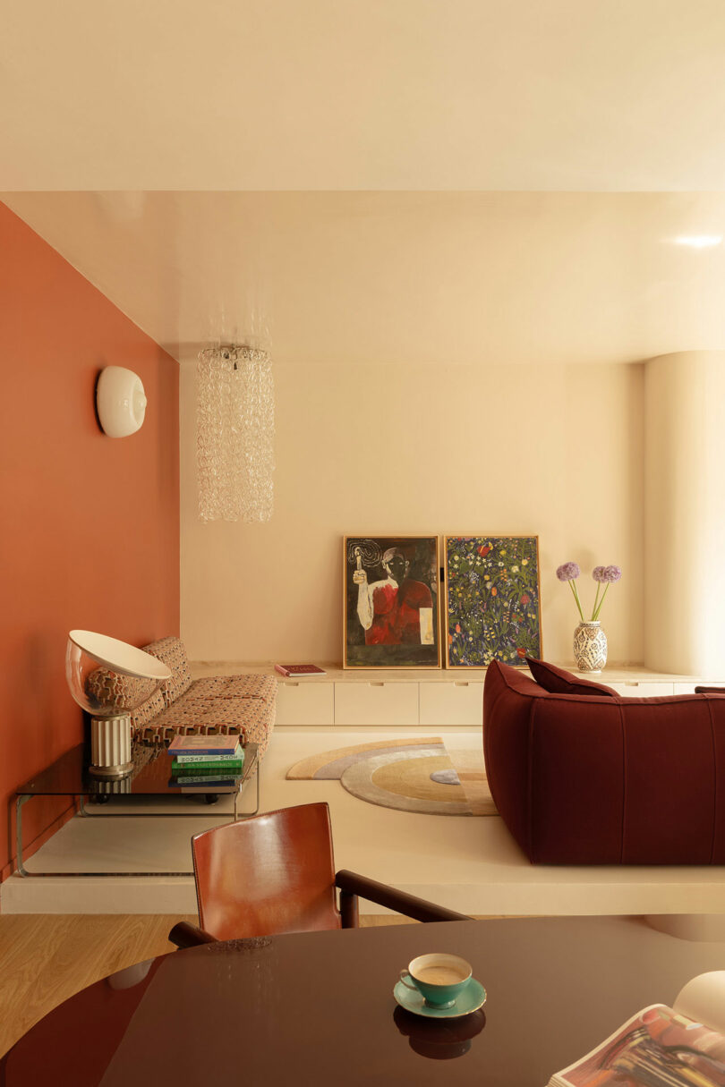 A modern living room with a red accent wall, sofa, a maroon chair, two framed artworks on the floor, decorative vases, and a glass table with a cup and magazine.